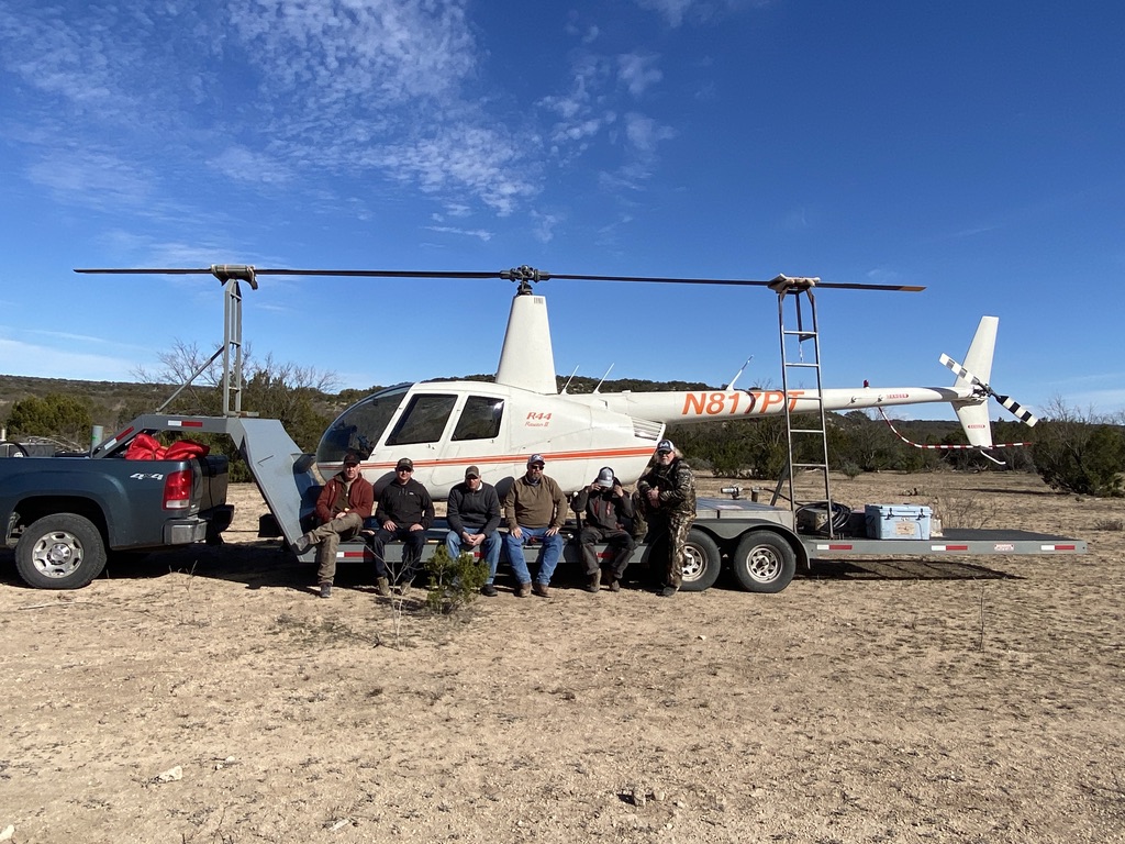 a dynamic, aerial view captures a helicopter hovering over a vast texas landscape, with well-equipped participants inside, focusing intently on their safety gear and protocols as they prepare for a thrilling hog hunting expedition.